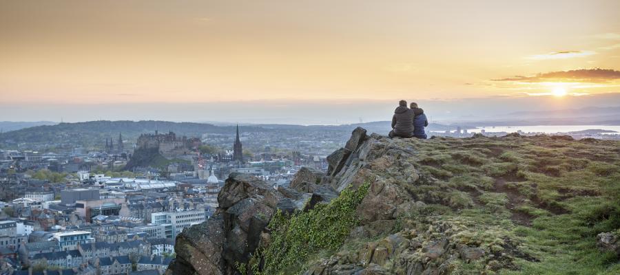 A pair of students sit on the edge of the Salisbury Crags, looking out over the city as the sun sets in the distance