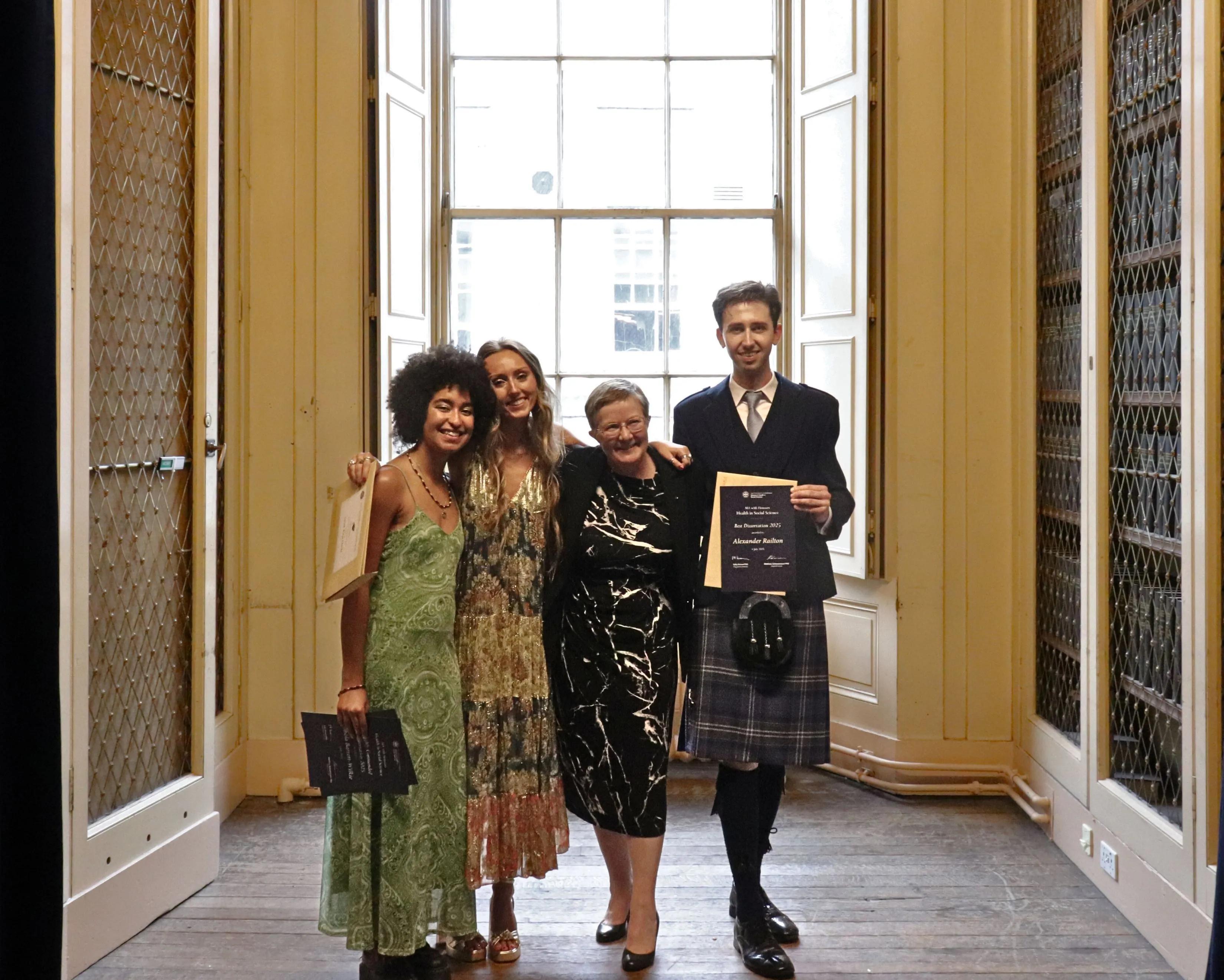 Three proud graduates pose with their programme director, hugging each other whilst holding up their awards