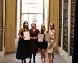 Three proud graduates pose with their course organiser, hugging each other whilst holding up their awards