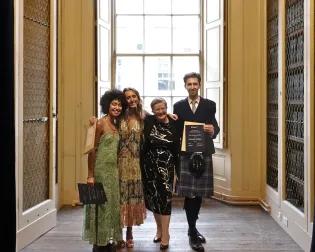 Three proud graduates pose with their programme director, hugging each other whilst holding up their awards