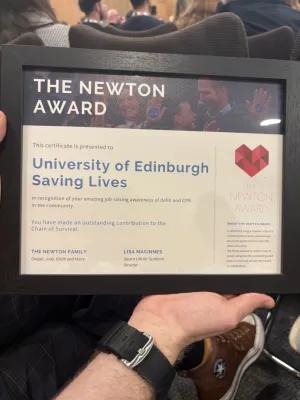 A hand holds up the framed Newton Award, awarded to the University of Edinburgh Saving Lives group